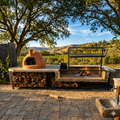 Outdoor patio kitchen setup with a terracotta wood-fired pizza oven on the left, a stone and metal grill station with wood storage in the center set under trees with a scenic rolling hills landscape in the background.