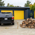 A black SUV parked in front of a modern garage with bright yellow doors, beside a 1/2 cord pile of almond firewood on a concrete driveway. Trees and sky are visible in the background.