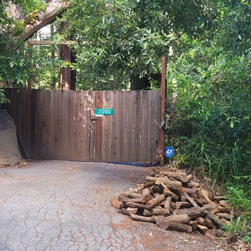 Wooden entry gate of home in Scott's Valley Ca with a pile of almond firewood logs in driveway surrounded by trees.
