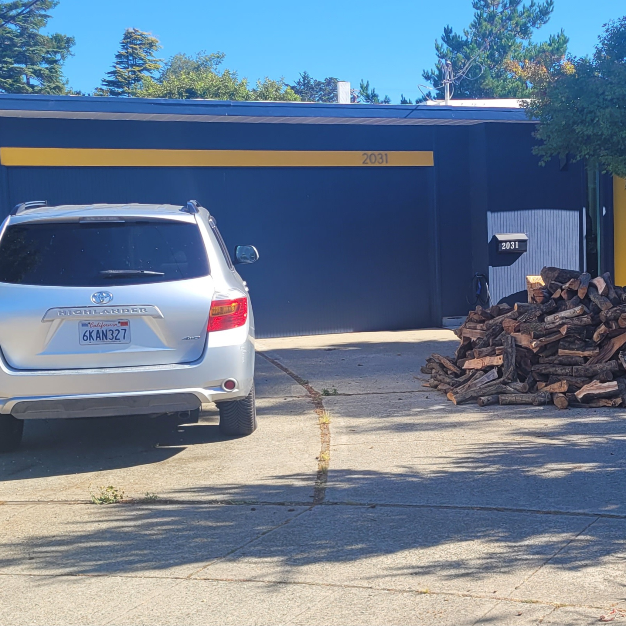 Silver car parked next to 1/3 cord of almond firewood in front of a blue house in San Mateo CA.