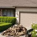 1/4 cord almonf firewood logs sits on the driveway in front of a beige stucco house with a closed garage door, trimmed green hedges, and white flowers in the foreground.
