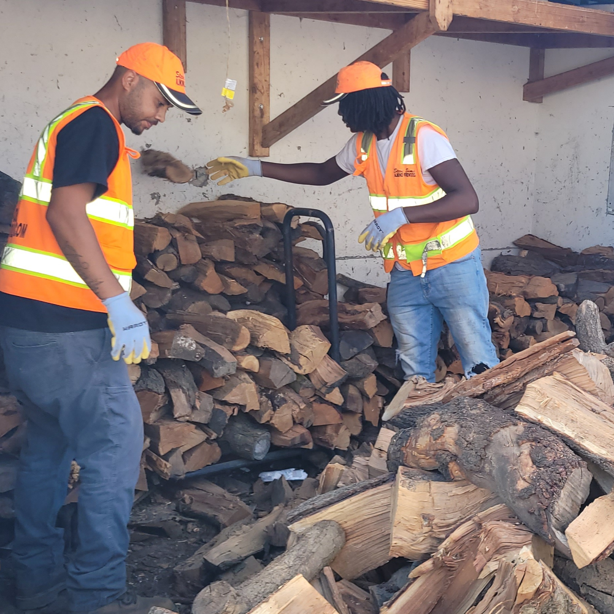 Two workers in safety vests and hats stacking 2 cords of almond firewood in a storage area for restaurant in San Carlos CA.