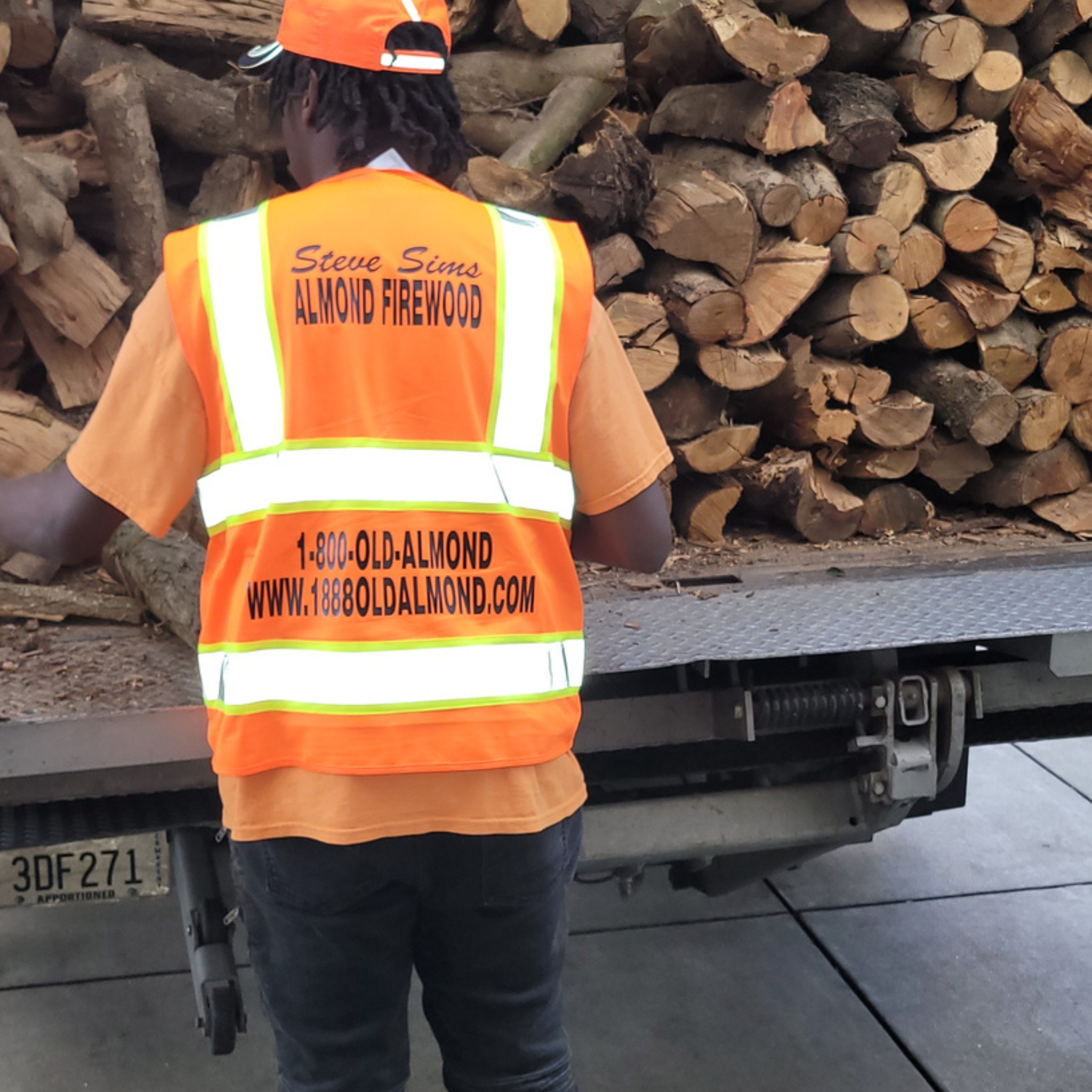 A worker wearing a bright orange high-visibility safety vest with reflective stripes and printed contact information for Steve Sims Almond Firewood, standing next to a truck loaded with stacked firewood logs.