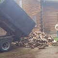 Truck unloading firewood logs onto a pile in front of a wooden cabin.