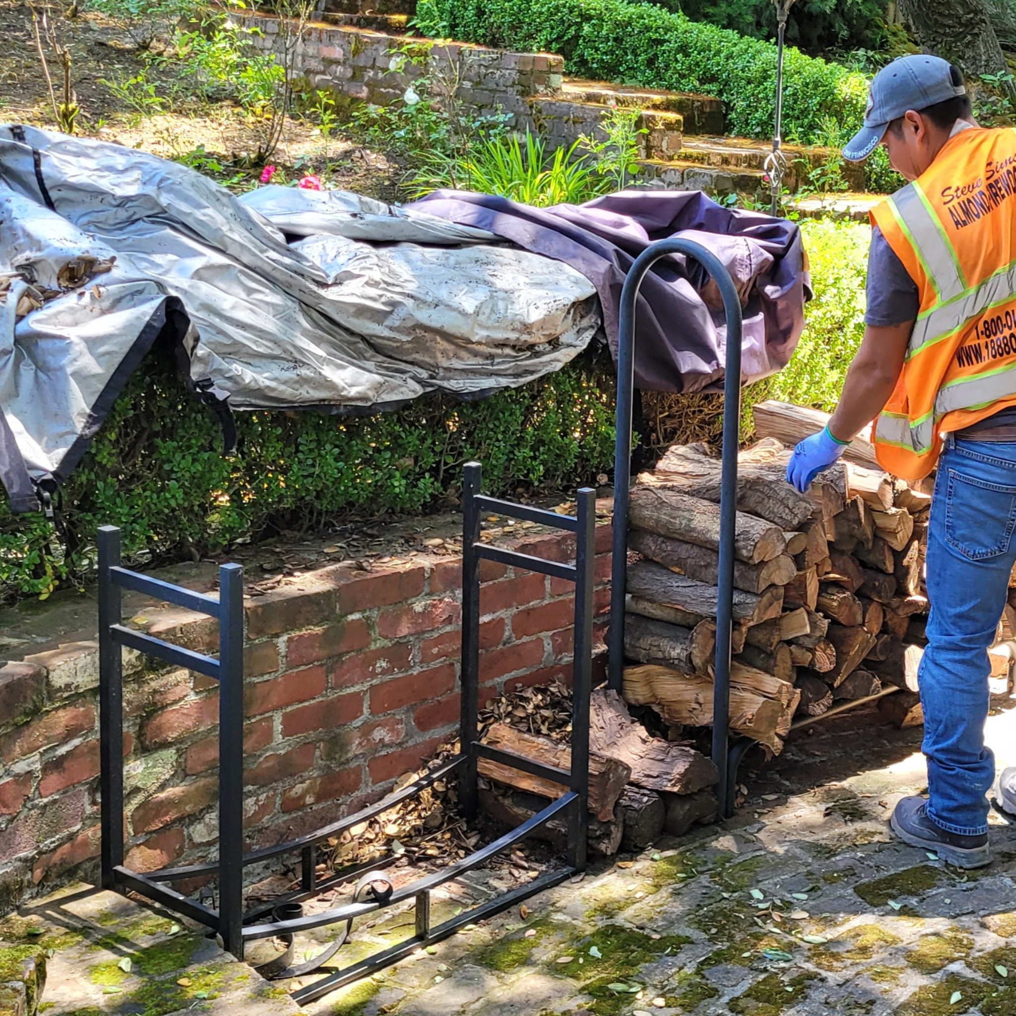 Person in an orange safety vest and blue cap stacking firewood o a metal rack on a stone patio.